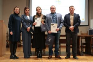 Grupa pięciu dorosłych osób stojących obok siebie na sali, na tle ekranu z wyświetloną prezentacją. Od lewej stoi kobieta o jasnej skórze, w ciemnej, długiej sukience lub tunice z golfem, w czarnych rajstopach i czarnych butach. Obok niej stoi kobieta o jasnej skórze w szarym, dwuczęściowym garniturze (marynarka i spodnie) oraz jasnej bluzce; trzyma w dłoni mikrofon. W środku stoi młoda kobieta o jasnej skórze, w czarnej, długiej sukience i czarnych butach; trzyma dyplom oraz złoty puchar. Czwarta osoba to mężczyzna o jasnej skórze w jasnoszarej marynarce, niebieskiej koszuli we wzór i dżinsach; trzyma dyplom oraz granatową torbę z białym napisem „UNIWERSYTET SZCZECIŃSKI” i logo. Piąta osoba to mężczyzna o jasnej skórze w ciemnej marynarce, beżowej koszuli we wzór i ciemnych spodniach, z rękami złożonymi z przodu. W tle widać ustawione stoły i krzesła oraz duży ekran projekcyjny; na ekranie jest widoczne logo i tekst, ale jest zbyt mało czytelny, by przepisać w całości. Na dole zdjęcia widnieje podpis: „fot. Magdalena Seredyńska/Uniwersytet Szczeciński”.