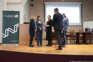 Scena w auli uniwersyteckiej z pięcioma dorosłymi osobami stojącymi na drewnianej scenie podczas wręczenia nagrody lub dyplomu. Dwie kobiety stoją po lewej stronie, zwrócone w stronę osoby odbierającej wyróżnienie; jedna ma jasne włosy do ramion i ciemną, dłuższą sukienkę lub płaszcz, druga ma ciemne włosy i niebieski garnitur. Pośrodku stoi młoda kobieta z długimi blond włosami, ubrana na czarno, trzymająca w dłoniach złoty puchar. Po prawej stoją dwaj mężczyźni; jeden bliżej aparatu w ciemnej marynarce i szarych spodniach, drugi w jasnoszarej marynarce, dżinsach i z długimi włosami związanymi z tyłu. W tle znajduje się długi stół i krzesła oraz ekran projekcyjny z wyświetloną prezentacją (tekstu nie da się tu wyraźnie odczytać). Po lewej stronie sceny stoi wysoki zielony roll-up z logo i czytelnym napisem: „UNIWERSYTET SZCZECIŃSKI”, „WYDZIAŁ NAUK ŚCISŁYCH I PRZYRODNICZYCH”, oraz na dole adres: „spr.usz.edu.pl”. W prawym dolnym rogu zdjęcia jest podpis: „fot. Magdalena Seredyńska/Uniwersytet Szczeciński”.