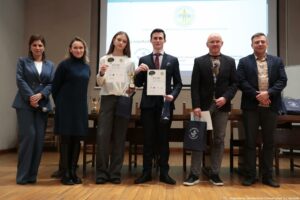 Grupa sześciu osób stojących w rzędzie na scenie w sali wykładowej, na tle dużego ekranu z wyświetloną prezentacją. Dwie osoby w środku trzymają dyplomy; kobieta w białej bluzce i szerokich, ciemnych spodniach trzyma kartkę z napisem „DYPLOM”, a mężczyzna w garniturze trzyma podobny dyplom oraz mały puchar. Na stole za nimi stoją dodatkowe puchary. Kilka osób trzyma granatowe torby prezentowe z białym napisem „Uniwersytet Szczeciński” i okrągłym logo. Po lewej stoją dwie kobiety: jedna w niebieskim garniturze z mikrofonem w dłoni, druga w ciemnej sukni z golfem. Po prawej stoją dwaj mężczyźni w marynarkach; jeden ma koszulkę z dużą, kolorową grafiką, drugi koszulę w jasny wzór. W prawym dolnym rogu zdjęcia widoczny jest podpis: „fot. Magdalena Seredyńska / Uniwersytet Szczeciński”.