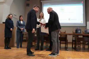 Scena wręczenia nagrody na sali wykładowej, z kilkoma osobami stojącymi na podwyższeniu przed dużym ekranem projekcyjnym. Na pierwszym planie dwóch mężczyzn podaje sobie ręce; mężczyzna po lewej ma ciemny garnitur i czarne buty, a mężczyzna po prawej ma czarną marynarkę, szare kraciaste spodnie i czarne sportowe buty z białą podeszwą. Między nimi, nieco z tyłu, stoi młoda kobieta z prostymi, brązowymi włosami do ramion, w białej bluzce i ciemnych spodniach; trzyma puchar oraz torbę prezentową. Po lewej stronie stoją dwie kobiety: jedna w ciemnej sukience z długim rękawem i czarnych wysokich butach, druga w niebieskim garniturze, trzymająca mikrofon. W tle znajdują się stoły i krzesła ustawione jak na konferencji oraz kilka granatowych toreb na stole. Na ekranie projekcyjnym widać stronę internetową z logo i tekstem po polsku, ale jest on w większości nieczytelny w tej rozdzielczości. W prawym dolnym rogu zdjęcia widnieje podpis: „fot. Magdalena Seredyńska/Uniwersytet Szczeciński”.