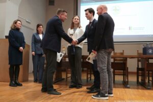 Grupa sześciu osób na scenie podczas uroczystego wydarzenia w sali wykładowej. Na środku widać uścisk dłoni dwóch mężczyzn w garniturach, jak podczas wręczania nagrody lub gratulacji. Obok stoi młoda kobieta w jasnej marynarce i spodniach, trzymająca puchar; na stole za nią stoją kolejne puchary. Po prawej stronie stoi łysy mężczyzna w czarnej marynarce i szarych spodniach, trzymający dyplom lub teczkę. Po lewej stronie stoją dwie kobiety: jedna w ciemnej, długiej sukience, druga w niebieskim garniturze. W tle jest duży ekran z wyświetloną stroną lub slajdem; widoczne są logotypy i napis „ZACHODNIOPOMORSKIE CENTRUM ONKOLOGII”. Na dole po prawej znajduje się podpis: „fot. Magdalena Seredyńska / Uniwersytet Szczeciński”.