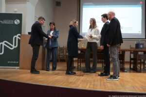 Scena wręczenia nagród na uczelnianej sali z sześcioma osobami stojącymi na podwyższeniu. Na środku kobieta w ciemnym płaszczu podaje dokument lub dyplom młodej kobiecie w białej koszuli i ciemnych spodniach; laureatka trzyma złoty puchar. Obok niej stoi młody mężczyzna w ciemnym garniturze, a po prawej mężczyzna w czarnym swetrze i jasnych, wzorzystych spodniach. Po lewej stronie mężczyzna w garniturze wręcza coś kobiecie w niebieskim garniturze, która trzyma torbę prezentową. W tle jest długi stół z kilkoma pucharami, krzesła oraz duży ekran z wyświetloną stroną internetową; z lewej stoi pionowy baner z napisem „UNIWERSYTET SZCZECIŃSKI” oraz „WYDZIAŁ NAUK ŚCISŁYCH I PRZYRODNICZYCH” i adresem „spr.usz.edu.pl”. W prawym dolnym rogu widoczny podpis: „fot. Magdalena Seredyńska/Uniwersytet Szczeciński”.