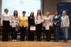 Grupa ośmiu kobiet stojących w rzędzie na scenie podczas uroczystości, na tle ekranu z prezentacją i dwóch granatowych ścianek z logo „Uniwersytet Szczeciński”. Kilka osób trzyma dyplomy; jedna osoba trzyma mikrofon, a przy niektórych widać ciemne torby upominkowe. Od lewej: Kobieta w białej koszuli i czarnych spodniach, blond włosy, okulary. Kobieta w jasnym swetrze, kręcone jasnobrązowe włosy; trzyma dyplom. Kobieta w żółtym żakiecie, krótkie czerwone włosy. Kobieta w czarnym stroju, długie ciemne włosy; trzyma jasną teczkę lub kartkę. Kobieta w jasnym golfie i ciemnych spodniach, długie jasnobrązowe włosy; trzyma dyplom. Kobieta w jasnej bluzce z różowawymi rękawami i ciemnych spodniach; trzyma dyplom. Kobieta w granatowym garniturze, ciemne włosy; w dłoni mikrofon. Kobieta w jasnej marynarce, jasnej koszuli i jasnych jeansach, blond włosy. Na slajdzie w tle da się odczytać m.in. „Przy współudziale” oraz widoczne są logotypy, w tym Polskiego Towarzystwa Fizjoterapii Medycznej i Uniwersytetu Szczecińskiego. Na dole zdjęcia widnieje podpis: „fot. Magdalena Seredyńska/Uniwersytet Szczeciński”.