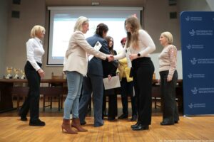 Grupa osób na scenie podczas uroczystości wręczania dyplomów. W centrum kadru dwie kobiety podają sobie ręce. Kobieta po lewej ma jasne blond włosy do ramion, jasną marynarkę i niebieskie dżinsy; stoi bokiem. Kobieta po prawej ma długie jasnobrązowe włosy, jasną bluzkę i czarne spodnie; w drugiej dłoni trzyma kartkę wyglądającą jak dyplom. Po bokach stoją kolejne osoby, również w stroju formalnym. Po lewej w tle widać stół z kilkoma złotymi pucharami. Z tyłu znajduje się duży ekran projekcyjny. Po prawej stronie kadru stoi niebieski roll-up z powtarzającym się napisem „UNIWERSYTET SZCZECIŃSKI” oraz logo „US”. Podłoga jest drewniana, a sala wygląda jak aula lub sala konferencyjna.