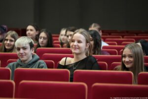 Grupa nastolatków siedzących na czerwonych fotelach w sali audytoryjnej. Na pierwszym planie są trzy osoby: po lewej chłopak o jasnych, krótko obciętych włosach w szarej bluzie z kapturem, pośrodku dziewczyna z jasnymi włosami związanymi z tyłu, w czarnym topie na ramiączkach i z naszyjnikiem, po prawej dziewczyna o długich, prostych włosach w jasnobrązowym kolorze. Wszyscy mają jasną skórę i delikatnie się uśmiechają. W tle widać więcej młodych osób siedzących w kolejnych rzędach; ostrość jest mniejsza. W prawym dolnym rogu jest mały napis: „fot. Magdalena Seredyńska/Uniwersytet Szczeciński”.