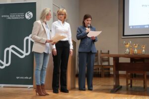 Trzy kobiety stojące na scenie podczas wydarzenia na Uniwersytecie Szczecińskim. Po lewej stronie widać roll-up z napisem: „UNIWERSYTET SZCZECIŃSKI”, „WYDZIAŁ NAUK ŚCISŁYCH I PRZYRODNICZYCH” oraz adres „spr.usz.edu.pl”, obok jest duże logo. Kobieta po lewej ma jasną karnację, jasne blond włosy do ramion, jasną marynarkę, jasnoniebieską koszulę, jasne dżinsy i brązowe botki na obcasie; trzyma teczkę z dokumentami. Kobieta pośrodku ma jasną karnację, blond włosy do ramion, okulary, białą koszulę i czarne spodnie; stoi przy mównicy. Kobieta po prawej ma jasną karnację i ciemne włosy upięte z tyłu; jest ubrana w granatowy garnitur i trzyma mikrofon oraz kartkę, z której czyta. Po prawej stronie stoi stół z trzema pucharami. W tle widać krzesła i fragment ekranu/projektora z uciętym tekstem zaczynającym się od „Przy w…”. Na dole zdjęcia jest podpis: „fot. Magdalena Seredyńska/Uniwersytet Szczeciński”.
