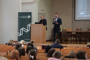 Aula wykładowa podczas wydarzenia akademickiego, z dwoma mężczyznami na scenie i publicznością na widowni. Po lewej stronie sceny stoi mównica z jasnego drewna, a za nią mężczyzna w ciemnym garniturze i czerwonym krawacie. Obok, po prawej, stoi drugi mężczyzna w ciemnym garniturze i białej koszuli, trzyma mikrofon w jednej dłoni i kartkę lub notatnik w drugiej, jakby prowadził zapowiedź lub przemówienie. Za mównicą widoczny jest pionowy baner w kolorze ciemnozielonym z białymi napisami: „UNIWERSYTET SZCZECIŃSKI” oraz „WYDZIAŁ NAUK ŚCISŁYCH I PRZYRODNICZYCH”, a także adres „spr.usz.edu.pl” i graficzny motyw przypominający falę lub wstęgę. Po prawej stronie sceny stoi długi stół z krzesłami; na stole ustawionych jest kilka pucharów (złote i srebrne, na ciemnych podstawach). W prawym górnym rogu jest fragment ekranu projekcyjnego z widocznym interfejsem komputera i listą opcji w języku polskim. Na pierwszym planie widać tyły głów publiczności siedzącej w drewnianych ławkach; dominują kobiety, różne fryzury i kolory włosów. W prawym dolnym rogu jest podpis fotograficzny: „fot. Magdalena Seredyńska/Uniwersytet Szczeciński”.