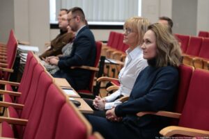 Rząd czerwonych foteli w sali konferencyjnej z kilkoma siedzącymi osobami, ujęty z boku pod kątem. Na pierwszym planie siedzą dwie kobiety zwrócone w prawą stronę, jakby słuchały prelegenta. Kobieta bliżej aparatu ma jasną cerę, falowane jasnobrązowe włosy do ramion i ma na sobie ciemny sweter. Obok niej siedzi kobieta o jasnej cerze z blond włosami z grzywką, w okularach, ubrana w białą koszulę. Obie mają dłonie splecione na kolanach. W dalszych rzędach siedzi kilka osób, w tym mężczyzna w granatowej marynarce i okularach oraz inni uczestnicy, częściowo rozmyci. Fotele są tapicerowane na czerwono z jasnymi drewnianymi podłokietnikami; między rzędami widać wąskie stoliki z notesami lub kartkami. Na dole po prawej widać mały podpis: „fot. Magdalena Seredyńska/Uniwersytet Szczeciński”.