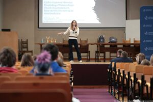 Sala wykładowa z audytorium, scena na podwyższeniu i osoba przemawiająca do publiczności z mikrofonem. Na pierwszym planie widać tyły głów osób siedzących na widowni oraz rzędy drewnianych siedzeń. Osoba na scenie stoi na środku, ma jasne, kręcone włosy do ramion i jasną karnację. Ubrana jest w jasny sweter z ciemnymi pasami przy mankietach i w okolicy talii oraz ciemne spodnie; ma jasne buty. Jedna ręka trzyma mikrofon przy ustach, druga jest wyciągnięta w bok w geście. Za nią stoi długi stół z kilkoma krzesłami. Na stole po lewej stronie są trzy złote puchary i butelka wody. Po prawej stronie leżą dwie ciemne torby z jasnym logo. Na ścianie wisi duży ekran z prezentacją w języku polskim. Czytelny tytuł slajdu: „Czyli o wyprawie Sławosza Uznańskiego‑Wiśniewskiego i trzech eksperymentach, które nieznacznie wpłyną na świat medycyny”. W prawym dolnym rogu zdjęcia jest podpis: „fot. Magdalena Seredyńska / Uniwersytet Szczeciński”. Po prawej stronie sceny stoi wysoki niebieski baner z napisem „Uniwersytet Szczeciński” i powtarzającym się logo.