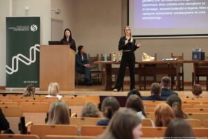 Aula wykładowa z publicznością siedzącą w drewnianych ławkach, z przodu scena z mównicą, stołem i ekranem projekcyjnym. Na scenie stoją dwie kobiety. Po prawej stronie stoi blondynka w okularach, ubrana na czarno (krótka marynarka i spodnie), trzyma mikrofon i pilot lub wskaźnik, wygląda na prowadzącą prezentację. Po lewej stronie, za drewnianą mównicą, stoi kobieta z długimi ciemnymi włosami w ciemnym stroju, obok niej na krześle siedzi kolejna osoba w garniturze, zwrócona w stronę mównicy. Po lewej stronie sceny stoi zielony roll-up z białym logo i napisem: „UNIWERSYTET SZCZECIŃSKI WYDZIAŁ NAUK ŚCISŁYCH I PRZYRODNICZYCH” oraz adresem „spr.usz.edu.pl”. Na stole stoją dwie złote statuetki/puchary, butelka wody i kilka przedmiotów, w tym coś przypominającego model anatomiczny ucha. Po prawej stronie leży torba lub pudełko z materiałami. Na ekranie projekcyjnym widać fragment slajdu w języku polskim o tematyce związanej z uchem (tekst jest ucięty i nie w całości czytelny). W prawym dolnym rogu zdjęcia widnieje podpis: „fot. Magdalena Seredyńska/Uniwersytet Szczeciński”.