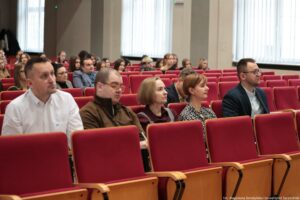 Sala wykładowa z rzędami czerwonych, tapicerowanych krzeseł audytoryjnych i grupą siedzących osób, ujęta z boku na poziomie siedzeń. Na pierwszym planie widać cztery osoby w jednym rzędzie: mężczyznę w białej koszuli, mężczyznę w brązowym swetrze z krótkimi włosami i okularami, kobietę z jasnymi, krótkimi włosami w ciemnym stroju oraz kobietę z krótkimi rudymi włosami w czarno-białym wzorzystym stroju. Po prawej stronie tego rzędu siedzi jeszcze mężczyzna w granatowej marynarce i okularach. Wszyscy są zwróceni w tę samą stronę, jakby słuchali prezentacji lub wykładu. W tle widać wiele innych osób rozproszonych po kolejnych rzędach, z dużymi odstępami między zajętymi miejscami. Większość ma neutralny lub skupiony wyraz twarzy. Po lewej stronie w głębi stoją wieszaki z okryciami. Na ścianie po prawej są wysokie okna z pionowymi żaluzjami, wpuszczające jasne światło dzienne. W prawym dolnym rogu jest drobny napis: „fot. Magdalena Seredyńska / Uniwersytet Szczeciński”.