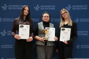 Trzy osoby pozujące do zdjęcia na tle ścianki z powtarzającym się napisem „UNIWERSYTET SZCZECIŃSKI” i logo uczelni. Po lewej stoi młoda kobieta o jasnej skórze, z długimi, prostymi, ciemnymi włosami. Ma na sobie ciemną, bordową koszulę i czarne spodnie. Trzyma w rękach dyplom. W środku stoi starsza kobieta o jasnej skórze, z ciemnymi włosami upiętymi do tyłu, w okularach. Ma na sobie czarny golf i szarą, bezrękawną kamizelkę oraz szare spodnie. Trzyma puchar w kolorze złotym oraz dyplom. Po prawej stoi młoda kobieta o jasnej skórze, z długimi, prostymi, jasnymi włosami, w dużych czarnych okularach. Ma na sobie czarny strój (marynarka i spodnie) oraz czarną bluzkę. Trzyma dyplom. Na dyplomach wyraźnie widać duży napis „DYPLOM”. W prawym dolnym rogu zdjęcia widoczny jest podpis: „fot. Magdalena Seredyńska/Uniwersytet Szczeciński”.