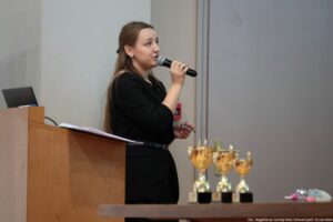 Kobieta przemawiająca do mikrofonu przy mównicy w sali konferencyjnej. Ma długie, proste brązowe włosy, jasną skórę i czarną sukienkę z paskiem w talii. Jest pokazana z boku, zwrócona w prawo, trzyma mikrofon przy ustach, a w drugiej dłoni ma mały przedmiot (prawdopodobnie pilot lub długopis). Na stole po prawej stronie stoi kilka złotych pucharów na czarnych podstawach. Na blacie widać też plastikową butelkę. W tle są jasne ściany i drzwi/panele. W prawym dolnym rogu jest podpis: „fot. Magdalena Seredyńska/Uniwersytet Szczeciński”.