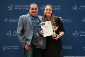 Dwie osoby pozujące do zdjęcia na tle granatowej ścianki z powtarzającym się napisem „UNIWERSYTET SZCZECIŃSKI” i logo „US”. Mężczyzna stoi po lewej, uśmiecha się, ma jasną cerę, ciemne włosy związane z tyłu, szarą marynarkę, granatową koszulę we wzory i dżinsy. Kobieta stoi po prawej, uśmiecha się, ma jasną cerę, długie jasnobrązowe włosy, czarną sukienkę i trzyma w dłoniach dyplom oraz złoty puchar na czarnej podstawie. Tekst widoczny na dyplomie: „DYPLOM” (pozostałe drobniejsze linie są nieczytelne w tej jakości). Dodatkowo widoczna jest ciemna torba z napisem „UNIWERSYTET SZCZECIŃSKI” i logo „US”. W prawym dolnym rogu zdjęcia jest podpis: „fot. Magdalena Seredyńska/Uniwersytet Szczeciński”.