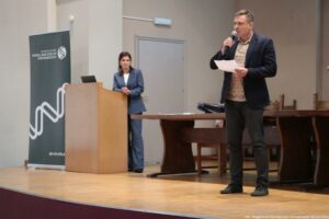 Scena w auli lub sali wykładowej z podwyższeniem, na którym stoi mężczyzna przemawiający do publiczności. Mężczyzna ma jasną cerę i krótkie jasne włosy, trzyma mikrofon przy ustach oraz kartkę papieru w drugiej dłoni. Jest ubrany w ciemną marynarkę, jasną koszulę we wzór, ciemne spodnie i ciemne buty. Po lewej stronie stoi drewniana mównica, a za nią kobieta o jasnej cerze i ciemnych, prostych włosach do okolic ramion. Ma na sobie niebieski garnitur i jasną bluzkę; dłonie ma złączone na wysokości brzucha. Na mównicy stoi otwarty laptop. Z lewej, obok mównicy, znajduje się pionowy roll-up z napisem: „UNIWERSYTET SZCZECIŃSKI WYDZIAŁ NAUK ŚCISŁYCH I PRZYRODNICZYCH” oraz okrągłym logo. Na dole roll-upu widoczny jest adres „sp.usz.edu.pl”. W tle widać duże drzwi oraz długi drewniany stół z krzesłami. W prawym dolnym rogu zdjęcia jest podpis: „fot. Magdalena Seredyńska/Uniwersytet Szczeciński”.