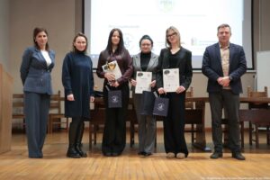 Grupa sześciu osób stojących w sali przed ekranem projekcyjnym, pozujących do zdjęcia podczas uroczystości wręczenia dyplomów. Na zdjęciu stoją cztery kobiety i jeden mężczyzna oraz jeszcze jedna kobieta; wszyscy ustawieni w jednym rzędzie. Dwie osoby w środku trzymają w dłoniach dyplomy; na dyplomach widoczny jest duży napis „DYPLOM”. Jedna z tych osób trzyma też złoty puchar. Dwie osoby mają również granatowe torby prezentowe z jasnym logo. Ubrania są formalne: marynarki, eleganckie spodnie i sukienka; dominują ciemne kolory (granat, czerń, szarość). Jedna kobieta ma jasne włosy i okulary, inna ma ciemne włosy upięte i okulary. Mężczyzna po prawej ma krótkie włosy i marynarkę. Tło: duży ekran z niewyraźnymi logotypami i tekstem, a przed nim długi stół i krzesła. Podłoga jest drewniana. W prawym dolnym rogu widoczny podpis fotograficzny: „fot. Magdalena Seredyńska / Uniwersytet Szczeciński”.