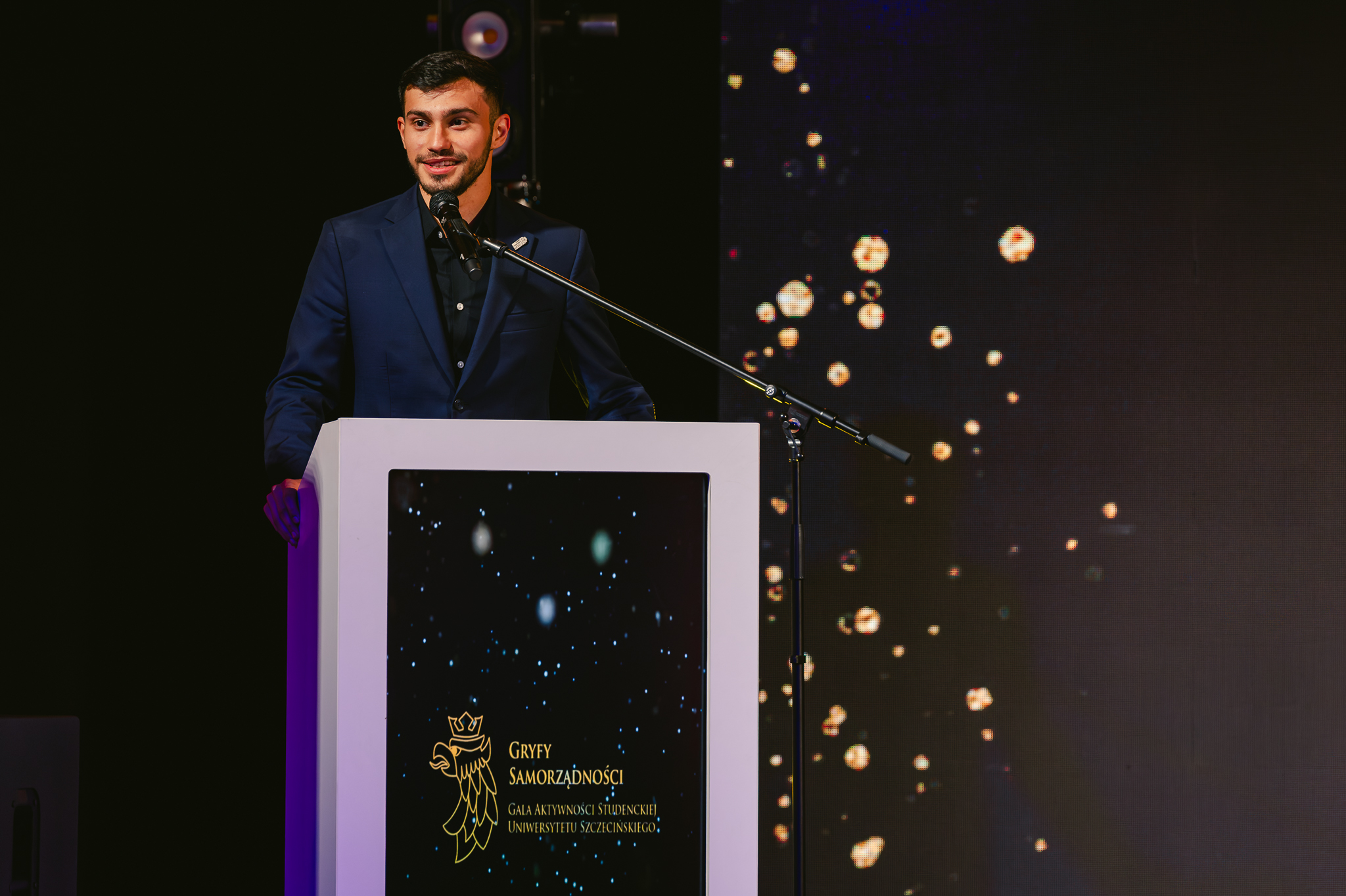 Mężczyzna w granatowym garniturze stoi za białą mównicą i przemawia do mikrofonu na statywie. Ma krótkie, ciemne włosy, krótki zarost i jasną do średniej karnację; jest lekko uśmiechnięty. Tło jest ciemne; po prawej stronie widać rozproszone, jasne punkty przypominające świetlne drobinki.