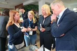 Grupa czterech osób rozmawiających przy stanowisku pokazowym w pomieszczeniu przypominającym targi lub wydarzenie uczelniane. Na pierwszym planie po lewej stoi kobieta o jasnej cerze i długich rudawych włosach, w okularach, w granatowym swetrze na białej koszuli; gestykuluje podczas rozmowy i trzyma niewielkie urządzenie wyglądające jak ręczne narzędzie lub prototyp (biało-niebieskie). Obok niej stoi kobieta o jasnej cerze i blond włosach związanych z tyłu, w okularach, w granatowej koszulce polo z logo oraz identyfikatorem na smyczy; w dłoni trzyma butelkę wody i słucha. Po prawej znajduje się kobieta o jasnej cerze i blond włosach do ramion, w ciemnej marynarce; trzyma w rękach ulotkę lub broszurę i jest zwrócona w stronę prezentującej. Najbardziej po prawej stoi mężczyzna o jasnej cerze i krótkich siwych włosach, w granatowej marynarce i białej koszuli; ma rękę przy twarzy w geście skupienia i też słucha.. Na dole po prawej jest podpis fotograficzny: „fot. Magdalena Seredyńska/Uniwersytet Szczeciński”.