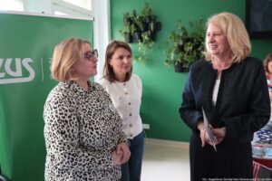 Trzy kobiety rozmawiające w pomieszczeniu, na tle zielonej ściany i zielonego roll-upu z widocznym fragmentem białego napisu „ZUS”. Po lewej stoi kobieta o jasnej skórze, blond włosach do brody i okularach, w białej bluzce w czarne cętki; ma na piersi przypięty identyfikator i mówi w stronę prawej. W środku stoi kobieta o jasnej skórze i brązowych włosach do ramion, w białym swetrze zapinanym na guziki; również ma przypinkę/identyfikator i patrzy na kobietę po prawej. Po prawej stoi kobieta o jasnej skórze i dłuższych blond włosach, w ciemnej marynarce i białej bluzce, uśmiecha się i trzyma w dłoniach kilka kartek lub broszur. Na ścianie w tle widać czarne doniczki z zielonymi roślinami. W prawym dolnym rogu jest podpis: „fot. Magdalena Seredyńska/Uniwersytet Szczeciński”.