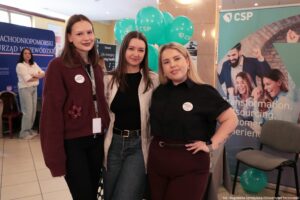 Trzy młode kobiety pozujące do zdjęcia na tle stoisk informacyjnych i zielonych balonów z logo „CSP”. Kobiety stoją blisko siebie w pomieszczeniu przypominającym halę lub korytarz podczas wydarzenia (np. targów). Lewa kobieta ma jasną skórę, proste brązowe włosy do ramion i ciemną szminkę. Ma na sobie bordowy sweter z ozdobą w kształcie kwiatu i przypiętą okrągłą plakietką; na szyi wisi identyfikator na smyczy. Dolna część stroju jest ciemna. Środkowa kobieta ma jasną skórę i długie, proste ciemnobrązowe włosy. Ma na sobie czarną bluzkę i jasną, cienką marynarkę w prążki oraz niebieskie dżinsy z paskiem. Prawa kobieta ma jasną skórę i długie blond włosy. Ma na sobie czarną koszulkę z okrągłą plakietką oraz bordowe spodnie z paskiem. Na lewym przedramieniu widać tatuaż, a na lewym nadgarstku smartwatch. W tle po prawej jest wysoki baner z napisem „CSP” u góry oraz fragmentami angielskich słów, m.in. „transformation” i „outsourcing”. Po lewej widać drugi baner z dużym napisem „ZACHODNIOPOMORSKI URZĄD WOJEWÓDZKI”. Na dole zdjęcia jest podpis: „fot. Magdalena Seredyńska/Uniwersytet Szczeciński”.