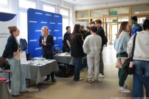 Stoisko informacyjne na korytarzu budynku, z kilkoma osobami rozmawiającymi przy dwóch stolikach i niebieskiej ściance reklamowej z logo „DECATHLON”. Pomieszczenie jest jasne, z oknami po lewej stronie i przeszklonymi drzwiami oraz ściankami działowymi z tyłu. Po lewej stronie stoi kobieta z włosami upiętymi w kok, w ciemnej marynarce i jasnych, szerokich spodniach jeansowych; jest zwrócona w stronę stolika. Obok, przy pierwszym stoliku, stoi starsza kobieta o blond włosach do ramion, w granatowym garniturze i jasnej koszuli; ma złożone dłonie i uśmiech. Na blacie stolika leżą rozłożone ulotki i materiały papierowe. Za stolikiem widać fragment białego roll-upu z napisem „Urząd Statystyczny w Szczecinie”. W centrum i po prawej stronie stoi grupa młodych osób (głównie w ciemnych kurtkach i bluzach) rozmawiających przy drugim stoliku z szarym obrusem. Jedna osoba stoi tyłem w jasnym dresie. Na stoliku są drobne przedmioty i papiery. W prawym tle stoi manekin ubrany w niebieską kamizelkę. Na dole po prawej jest podpis fotograficzny: „fot. Magdalena Seredyńska/Uniwersytet Szczeciński”.