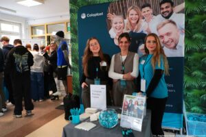 Stoisko targowe firmy Coloplast w holu budynku, z trzema kobietami stojącymi za stołem z materiałami promocyjnymi. Na stole leżą ulotki i wizytówki, stoi pojemnik z niebieskimi długopisami, szklana misa wypełniona niebieskimi cukierkami oraz stojak z broszurami; widoczny jest też napis „Coloplast Business Centre”. Za stoiskiem stoi duży baner z logo „Coloplast” i dużym zdjęciem uśmiechniętych osób. Kobiety mają identyfikatory na smyczach; dwie mają okulary, a jedna ma na sobie turkusową koszulkę. Po lewej stronie zdjęcia widać grupę ludzi stojących w kolejce lub rozmawiających przy innych stanowiskach oraz manekina w sportowym stroju (czapka, niebieska kamizelka, czarne spodenki). Na dole po prawej stronie znajduje się podpis: „fot. Magdalena Seredyńska / Uniwersytet Szczeciński”.