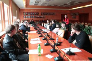 Sala konferencyjna z długim, błyszczącym drewnianym stołem i wieloma mikrofonami na podstawach ustawionymi wzdłuż blatu. Wokół stołu siedzi kilkanaście osób, głównie młodych dorosłych, ubranych na co dzień; część ma okulary. Na stole leżą kartki papieru, a na pierwszym planie stoi zielona butelka napoju. Po prawej stronie, bliżej końca stołu, stoi kobieta w czerwonym swetrze i okularach, zwrócona w stronę grupy, jak podczas prowadzenia spotkania. Pomieszczenie ma ciepłe, pomarańczowo-brązowe ściany; na ścianie z tyłu widać rzędy oprawionych portretów W prawym dolnym rogu widać mały podpis: „fot. Magdalena Seredyńska/Uniwersytet Szczeciński”.