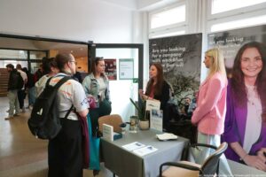 Stanowisko informacyjne w holu budynku, z szarym stołem i dużymi pionowymi bannerami reklamowymi w tle. Przy stole stoją cztery kobiety i rozmawiają; w tle po lewej stronie widać więcej osób w kolejce lub przechodzących przez otwarte drzwi. Na stole leżą ulotki i kartki, stoi mały stojak z materiałami oraz pudełko. Jest też mała roślina w doniczce i pojemnik z długopisami. W prawym dolnym rogu widać mały podpis: „fot. Magdalena Seredyńska/Uniwersytet Szczeciński”.