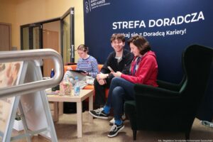 Strefa doradcza w budynku uczelni z trzema osobami siedzącymi na pomarańczowej kanapie przy małym stoliku, na tle granatowej ściany z dużym napisem. Na ścianie widoczny jest tekst: „STREFA DORADCZA” oraz niżej „Spotkaj się z Doradcą Kariery!”. W lewym górnym rogu ściany znajduje się logo i napis „AKADEMICKIE BIURO KARIER UNIWERSYTETU SZCZECIŃSKIEGO”. Po prawej stronie siedzi kobieta w czerwonej bluzie, dżinsach i czarnych butach sportowych; trzyma telefon i uśmiecha się, odwrócona lekko w stronę mężczyzny obok. Obok niej siedzi młody mężczyzna w ciemnej bluzie i czarnych spodniach, uśmiechnięty, z rękami opartymi na kolanach. Po lewej stronie na kanapie siedzi młoda kobieta w biało-granatowej bluzce w paski; pisze lub rysuje na podkładce/notesie, pochylona nad nim.