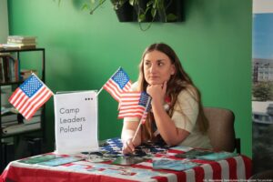 Stanowisko informacyjne z obrusem w amerykańskie barwy (czerwono-białe pasy i niebieskie pole z białymi gwiazdami). Na środku stołu stoi przezroczysta ramka z kartką z napisem „Camp Leaders Poland” oraz trzema kropkami w prawym górnym rogu. Na stole leżą rozłożone ulotki i broszury w różnych kolorach. Widoczne są dwie małe amerykańskie flagi na patyczkach: jedna po lewej stronie przy ramce, druga trzymana przy prawej stronie stołu. Po prawej stronie siedzi młoda kobieta o jasnej skórze i długich brązowych włosach, w jasnej koszulce. Opiera brodę na dłoni i ma neutralny, lekko zamyślony wyraz twarzy; wzrok skierowany w bok, poza kadr. W tle jest zielona ściana, fragment regału z książkami po lewej oraz wisząca doniczka z pnącą rośliną u góry. W prawym dolnym rogu widać mały podpis: „fot. Magdalena Seredyńska/Uniwersytet Szczeciński”.
