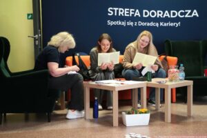 Strefa doradcza z trzema kobietami siedzącymi przy stolikach, na tle granatowej ściany z dużym białym napisem. Na ścianie widoczny tekst: “STREFA DORADCZA” oraz niżej “Spotkaj się z Doradcą Kariery!”. Po lewej stronie siedzi kobieta o jasnych, krótkich, kręconych włosach, ubrana na ciemno; pochylona, pisze lub notuje na kartce. Na środku siedzi młoda kobieta o prostych, brązowych włosach, w szarym ubraniu, trzyma i czyta kartkę. Po prawej siedzi młoda kobieta o długich, jasnych włosach i okularach, w jasnej kurtce; również trzyma i czyta kartkę. Siedzą na pomarańczowej kanapie, a po bokach stoją ciemnozielone fotele. Z przodu są dwa jasne, drewniane stoliki z rozłożonymi papierami i długopisami. Na prawym stoliku stoi słoik z kolorowymi cukierkami, butelka wody oraz puszka napoju. Na podłodze przy stolikach widać niebieską butelkę i białą donicę z kwiatami. Na dole zdjęcia leży kilka okrągłych przypinek lub żetonów. W prawym dolnym rogu widoczny podpis: “fot. Magdalena Seredyńska / Uniwersytet Szczeciński”.