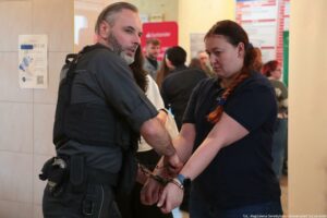 Dwójka dorosłych ludzi w centrum kadru, stojących w pomieszczeniu wśród innych osób. Mężczyzna po lewej ma jasną skórę, krótkie ciemne włosy z siwizną i siwą brodę; ma na sobie ciemny strój i kamizelkę z pasami oraz elementami wyposażenia przy pasie. Kobieta po prawej ma jasną skórę i długie rude włosy związane z tyłu; ma na sobie granatową koszulkę polo i opaskę/zegarek na lewym nadgarstku. Nadgarstki tych dwóch osób są połączone metalowymi kajdankami, a obie osoby trzymają ręce blisko siebie na wysokości pasa. W tle widać kilka rozmytych postaci.