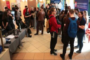 Tłum ludzi w holu budynku podczas wydarzenia informacyjnego z kilkoma stoiskami. Widać kilkanaście młodych dorosłych osób rozmawiających w grupach; część ma plecaki i kurtki. Po lewej stronie na stole z szarym obrusem leżą przedmioty przypominające broń długą i krótką oraz pałki; obok są kubki i papiery. W tle stoją roll-upy i tablice informacyjne, m.in. z napisem „STREFA DORADCZA” oraz duży baner „ZACHODNIOPOMORSKI URZĄD WOJEWÓDZKI”. Na dole po prawej jest podpis fotograficzny: „fot. Magdalena Seredyńska/Uniwersytet Szczeciński”.