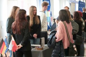 Grupa osób przy stoisku informacyjnym w jasnym wnętrzu, wydarzeniu uczelnianym. W centrum stoją trzy młode kobiety: jedna z długimi, jasnymi włosami w czarnej marynarce, obok kobieta z długimi ciemnoczerwonymi włosami w czarnym stroju oraz kobieta w okularach i szarej marynarce. Przed nimi znajduje się stół z szarym obrusem; na stole stoi miska z drobnymi słodyczami oraz pionowa stojąca kartka/informator. Po lewej stronie, na pierwszym planie, widać małe flagi na patyczkach: Unii Europejskiej, Niemiec i Polski. Po prawej stronie stoi kilka osób odwróconych plecami; najbardziej widoczna jest kobieta w różowym swetrze z wypukłymi kropkami, z torbą na ramieniu i trzymaną ciemną kurtką. W tle są banery reklamowe; na jednym widać napis „Erasmus” (reszta tekstu jest częściowo zasłonięta). W prawym dolnym rogu jest podpis: „fot. Magdalena Seredyńska/Uniwersytet Szczeciński”.