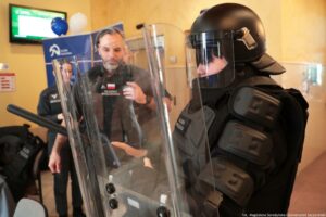 Funkcjonariusz w czarnym rynsztunku ochronnym i kasku z przezroczystą przyłbicą, stojący z profilu obok przezroczystej tarczy ochronnej. Na pierwszym planie po prawej stronie widać osobę w pełnym czarnym umundurowaniu ochronnym: kask, przyłbica, osłona twarzy, kamizelka i ochraniacze na ramionach. Przed nią znajduje się duża, przezroczysta tarcza z uchwytami i metalowymi elementami. W tle stoi mężczyzna w ciemnym ubraniu z naszywką z polską flagą i napisem „SŁUŻBA WIĘZIENNA”, a obok niego częściowo widoczna kolejna osoba w podobnym stroju. Pomieszczenie wygląda na wnętrze instytucji; w górnej lewej części jest ekran, a w tle widać balony i niebieski baner. W prawym dolnym rogu jest podpis zdjęcia: „fot. Magdalena Seredyńska / Uniwersytet Szczeciński”.