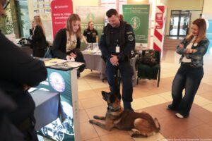 Stoisko informacyjne w holu, przy którym stoi umundurowany policjant z psem leżącym na podłodze. W centrum widać policjanta w granatowym mundurze z naszywką „POLICJA”; obok niego leży brązowy pies służbowy (wygląda jak owczarek) w pozycji „waruj”, z obrożą. Po lewej stronie młoda kobieta o jasnej cerze i długich rudawych włosach pochyla się nad blatem stoiska i przegląda materiały (kartki, broszury). Po prawej stoi kobieta o jasnej cerze i jasnobrązowych włosach, w dżinsach i kurtce dżinsowej, z założonymi rękami, uśmiechnięta.