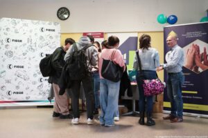 Grupa osób stojących przy stoisku informacyjnym w pomieszczeniu, obok dużych banerów reklamowych i zegara na ścianie. Kilka młodych osób z plecakami i torbami stoi w skupieniu przy stole, jakby rozmawiały z obsługą stoiska lub oglądały materiały. Po prawej stronie stoi łysy mężczyzna w okularach, w jasnej koszuli we wzory i jeansach; wygląda na osobę prowadzącą rozmowę lub prezentację.