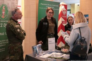 Stan informacyjny Straży Granicznej z trzema funkcjonariuszami za stołem oraz kobietą stojącą przed stoiskiem i rozmawiającą z nimi. Scena wygląda na wydarzenie w budynku (hala lub korytarz) z materiałami promocyjnymi na stole. Po lewej stronie stoi mężczyzna w mundurze moro z naszywką „POLAND” na rękawie; ma krótkie siwe włosy i okulary oparte na głowie. Na środku stoi kobieta w ciemnej kurtce służbowej i spodniach moro; ma długie jasnobrązowe włosy i okulary. Po prawej, za stołem, siedzi lub stoi niżej druga kobieta w czarnym umundurowaniu; ma jasne włosy do ramion. Przed stoiskiem stoi kobieta odwrócona tyłem w jasnym płaszczu, z ciemnym plecakiem; trzyma w ręku ulotkę lub broszurę. Na stole leżą liczne ulotki, broszury i foldery, stojak z materiałami, a także szklana misa z cukierkami. Na środku stołu stoi pionowa tabliczka z napisem „Straż Graniczna”.