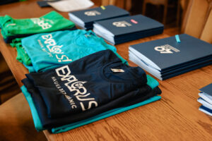 Ujęcie bokiem. Na drewnianym stole granatowe, niebieskie i zielone koszulki z napisem: explorUS! Nauka bez granic. Na drugim planie grantowe teczki z białym logiem 40-lecia Uniwersytetu Szczecińskiego.