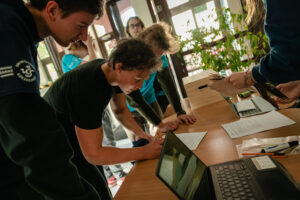 Korytarz. Pośrodku podłużny stół. Po lewej pochyleni nad stołem uczniowie, którzy wypełniają zadanie w grze „Naukowy Detektyw – Misja w Terenie”. W tle przeszklone wejście do budynku.