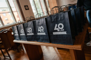 Ujęcie bokiem. Na drewnianym stole ustawione w pięciu rzędach granatowe papierowe torby z białym logiem 40-lecia Uniwersytetu Szczecińskiego, stanowiące nagrody w grze „Naukowy Detektyw – Misja w Terenie”.