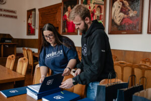 Dwoje organizatorów gry „Naukowy Detektyw – Misja w Terenie” przeglądają ułożone na drewnianym stole granatowe teczki z białym logiem 40-lecia Uniwersytetu Szczecińskiego, w których znajdują się certyfikaty uczestnictwa. W tle krzesła i na ścianie duże portrety rektorów ubranych w gronostaje.