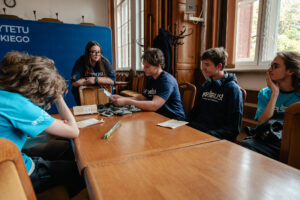 Przy drewnianym stole siedzą uczestnicy gry „Naukowy Detektyw – Misja w Terenie” i omawiają zadania z organizatorką. Na stole leżą Dzienniki badawcze używane podczas zadań. W tle okna i stojący wieszak z kurtkami.