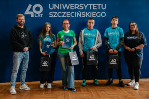 Czworo ubranych w niebieskie i zielone koszulki z napisem: „explorUS! Nauka bez granic” uczestników gry „Naukowy Detektyw – Misja w Terenie” z nagrodami (granatowe teczki i torby z białym logiem 40-lecia Uniwersytetu Szczecińskiego). Stoją z dwojgiem organizatorów na tle granatowej ścianki z białym logiem 40-lecia US.