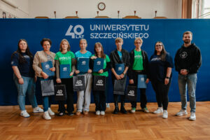 Sześcioro ubranych w niebieskie i zielone koszulki z napisem: „explorUS! Nauka bez granic” uczestników gry „Naukowy Detektyw – Misja w Terenie” z nagrodami (granatowe teczki i torby z białym logiem 40-lecia Uniwersytetu Szczecińskiego). Stoją z trojgiem organizatorów na tle granatowej ścianki z białym logiem 40-lecia US.