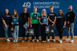 Pięcioro ubranych w grantowe i zielone koszulki z napisem: „explorUS! Nauka bez granic” uczestników gry „Naukowy Detektyw – Misja w Terenie” z nagrodami (granatowe teczki i torby z białym logiem 40-lecia Uniwersytetu Szczecińskiego). Stoją z trojgiem organizatorów na tle granatowej ścianki z białym logiem 40-lecia US.