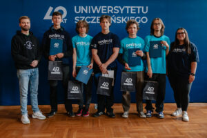 Pięcioro ubranych w grantowe i zielone koszulki z napisem: „explorUS! Nauka bez granic” uczestników gry „Naukowy Detektyw – Misja w Terenie” z nagrodami (granatowe teczki i torby z białym logiem 40-lecia Uniwersytetu Szczecińskiego). Stoją z dwojgiem organizatorów na tle granatowej ścianki z białym logiem 40-lecia US.