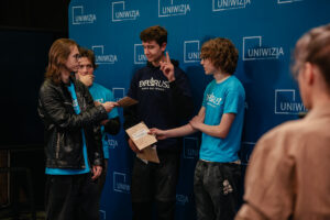 Ujęcie bokiem. Czworo ubranych w grantowe i niebieskie koszulki z napisem: „explorUS! Nauka bez granic” uczestników gry „Naukowy Detektyw – Misja w Terenie” stoi na tle granatowej ścianki z białymi logami UNIwizji – Telewizji Uniwersytetu Szczecińskiego.