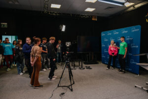 Po prawej dwoje uczestników gry „Naukowy Detektyw – Misja w Terenie” stoi na tle granatowej ścianki z białymi logami UNIwizji – Telewizji Uniwersytetu Szczecińskiego. Po lewej kamera i lampa na statywie, operator i kilkoro obserwatorów, w tym innych uczestników zabawy.