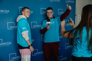 Dwóch ubranych w niebieskie koszulki z napisem: „explorUS! Nauka bez granic” uczestników gry „Naukowy Detektyw – Misja w Terenie” stoi na tle granatowej ścianki z białymi logami UNIwizji – Telewizji Uniwersytetu Szczecińskiego. Chłopak po lewej trzyma w jednej ręce mikrofon, drugą pokazuje kciuk uniesiony w górę. Tyłem do kadru dziewczyna w takiej samej koszulce robiąca im zdjęcie telefonem.